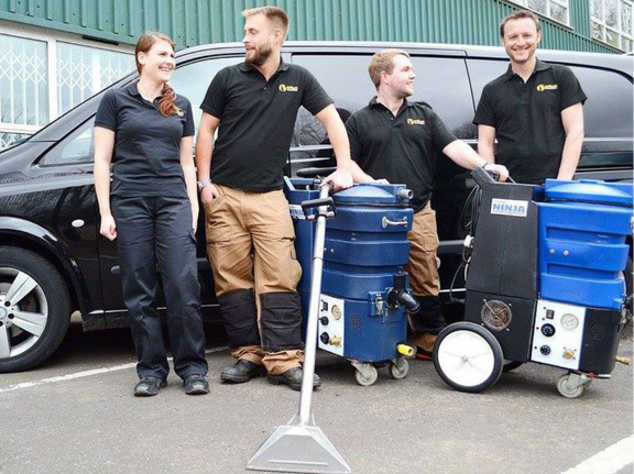 This is a photo of Blackheath Carpet Cleaning carpet cleaners (three men and one woman) standing in fromt of their black van, with two steam cleaning carpet machines next to them